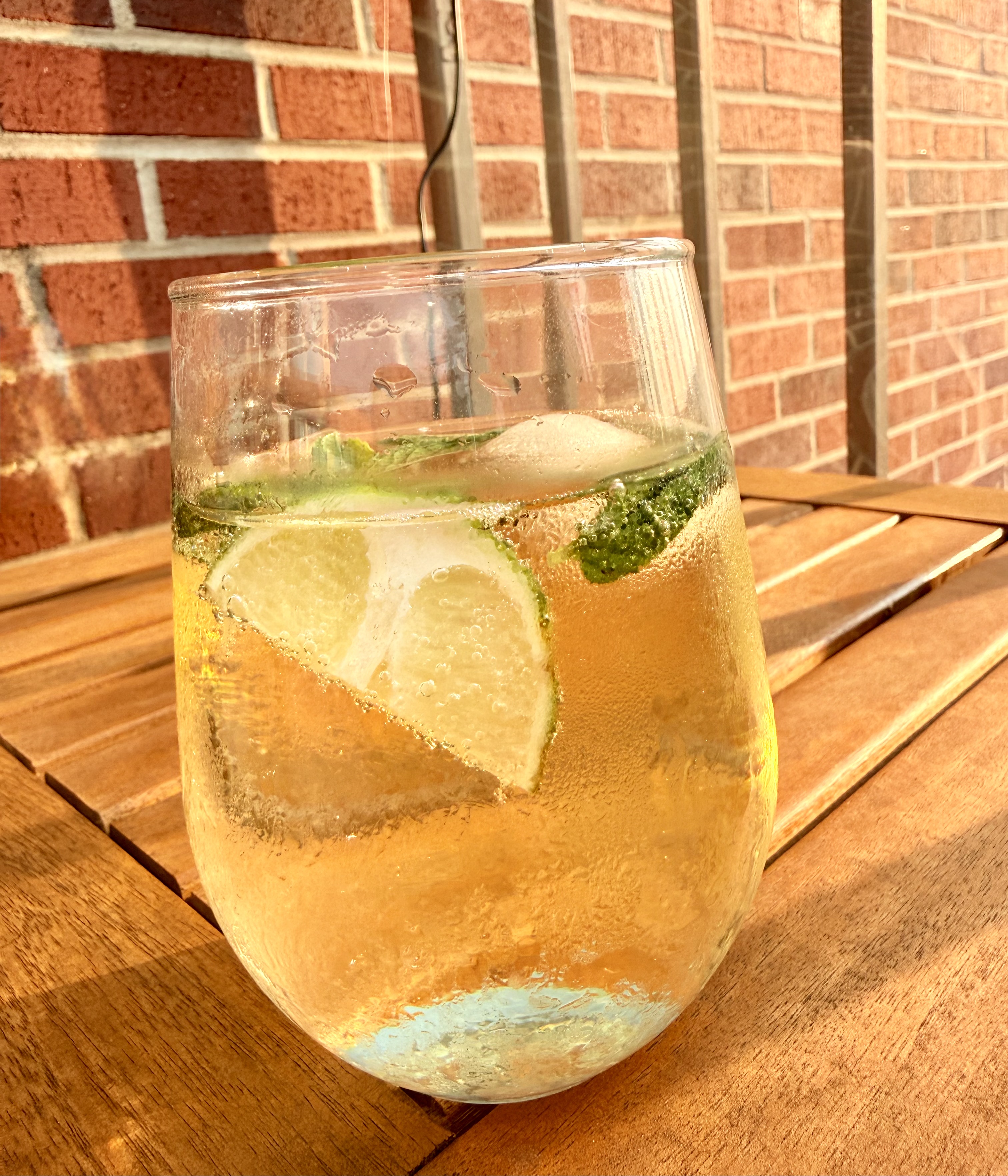 A Hugo Spritz on a wooden table, lime and mint suspended in the glass against a brick wall