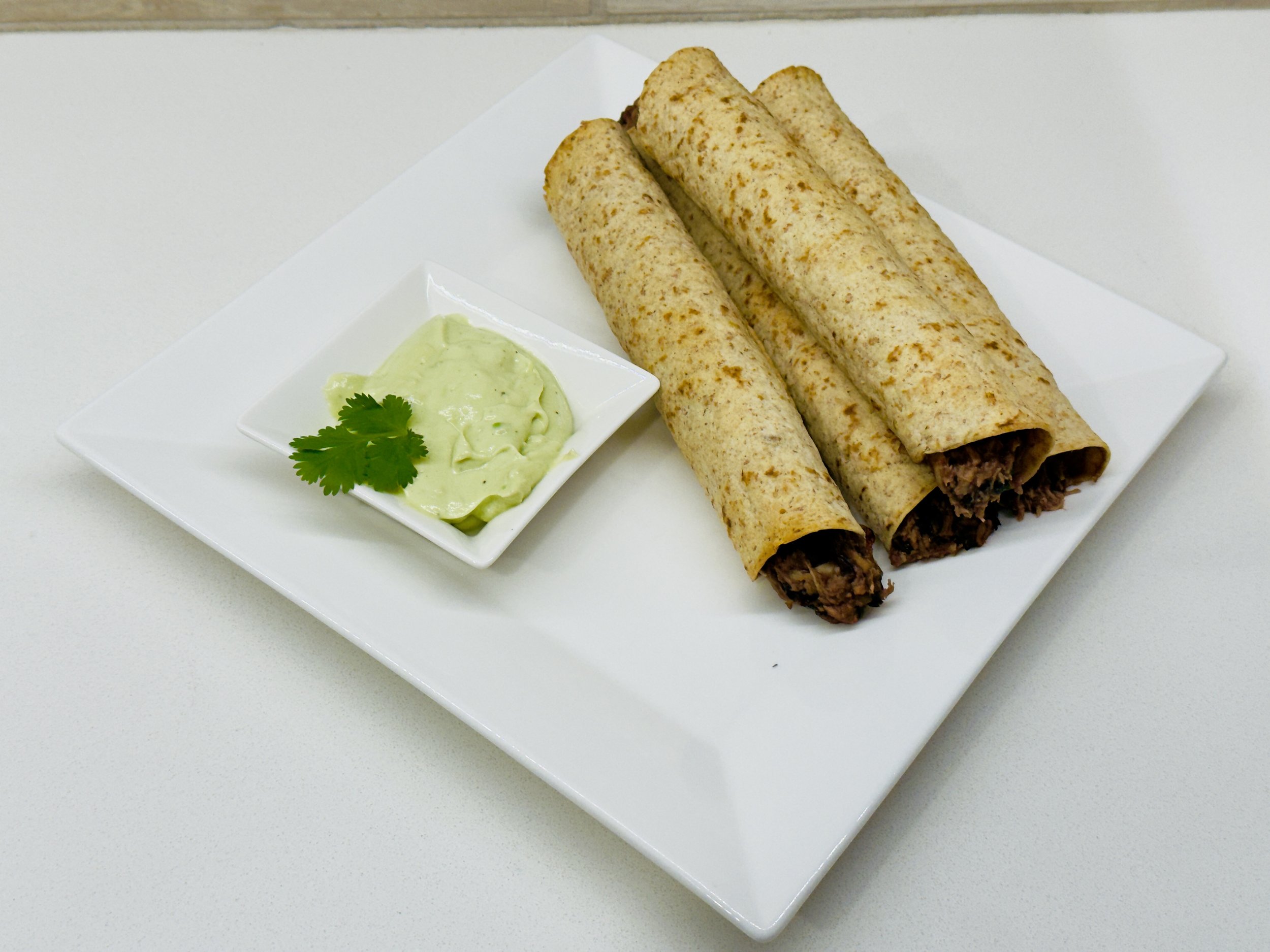 A plate of crispy taquitos with a small bowl of avocado-lime sauce for dipping